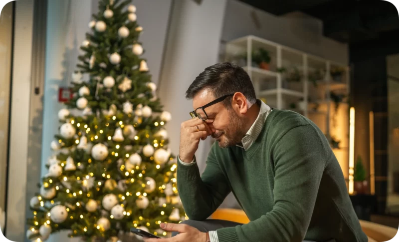 A man with glasses appears stressed or sad, holding his phone near a festive Christmas tree, possibly affected by seasonal affective disorder.