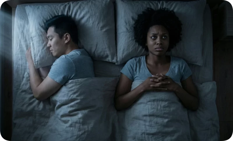 Self-Doubt in Relationships: 5 Ways to Rebuild Self-Trust 1 Anxious woman wide awake in bed, clasping hands, next to a peacefully sleeping man; visualizing self-doubt in relationships.