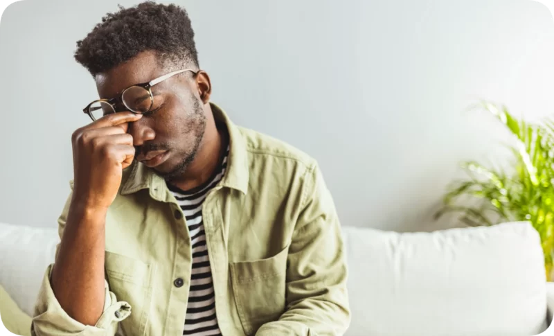 A young Black man with glasses pinches his nose, eyes closed, suggesting stress or fatigue. Reflects anxiety as a signal.