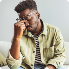 Young Black man pinching bridge of nose, eyes closed, on couch. He's experiencing distress, recognizing anxiety as a signal.