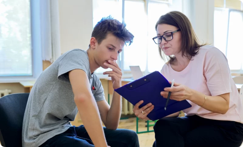 Female therapist with glasses and a young male client sit together reviewing a clipboard during a types of therapy session in a bright room