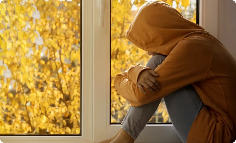 Sad teen in orange hoodie sitting alone by window hugging knees — sign teen therapy is needed