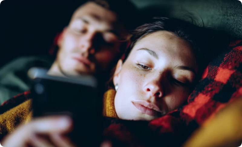 Couple lying together in bed at night, woman scrolling her phone with tired eyes while man rests in background, showing the impact of late-night doomscrolling