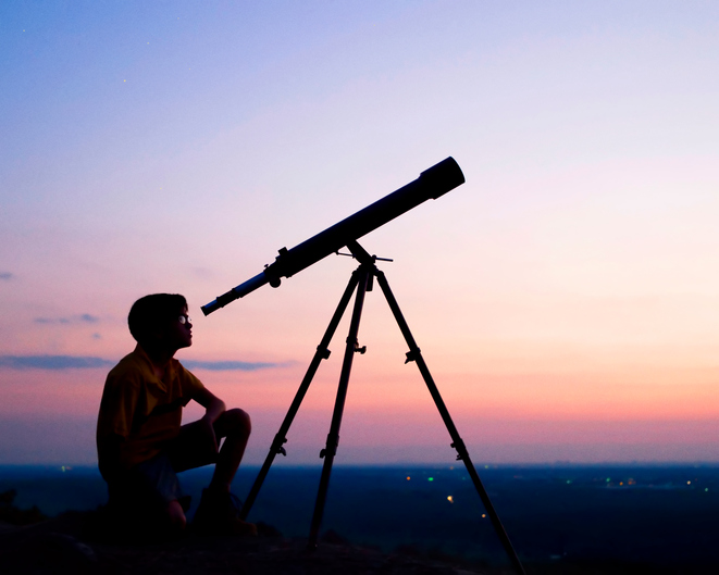 Silhouette of a boy looking through a telescope at sunrise.