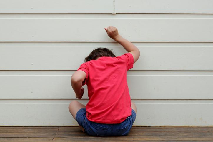 Child in red shirt, angry, facing away and hitting wall