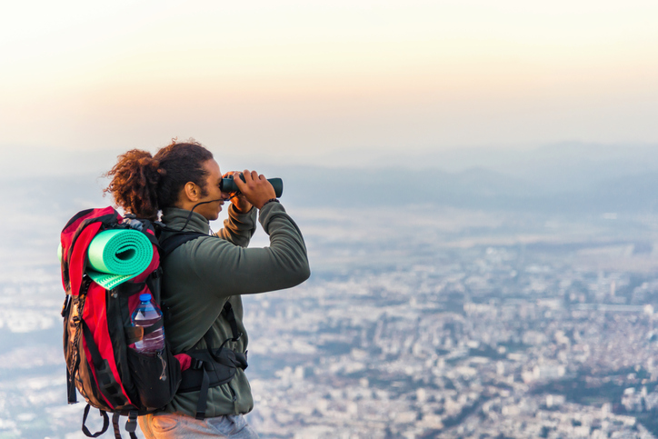 Man standing at the top of a hill and looking at view though binoculars at sunset.