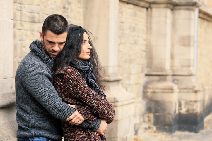 A man hugs a woman from behind. They both look upset.