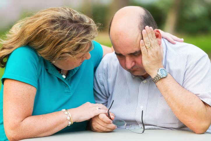 A woman in a blue shirt consoles a balding man who looks sadly at his glasses.