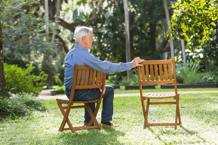 An elderly man sits outdoors next to an empty lawn chair.