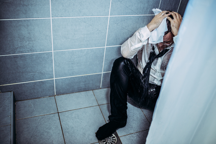 A man in a suit sits on the ground, covering his head with his hands. He is very wet.