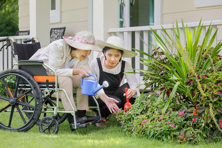 A senior woman in a wheelchair waters her garden. Her younger daughter kneels beside her to help.