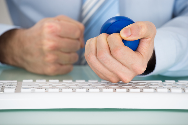 A businessman sits in front of a keyboard, making a fist with one hand and squeezing a stress ball in the other.