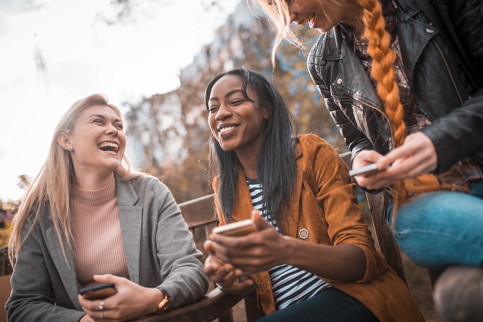 Three friends laughing and talking on bench in the park.