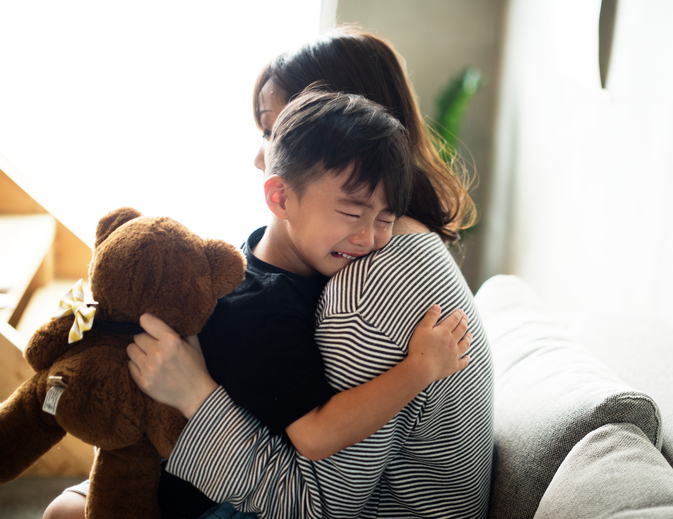 A mother hugs her crying son while holding his teddy bear for him.