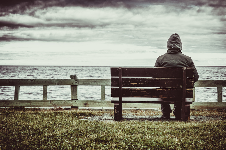 A man in a rain jacket sits on a bench overlooking a cloudy sea.