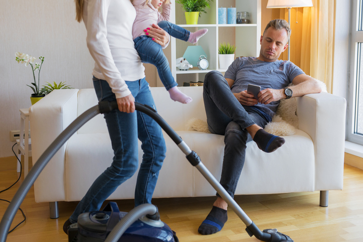 A wife vacuums and carries a toddler while her husband plays on his phone.