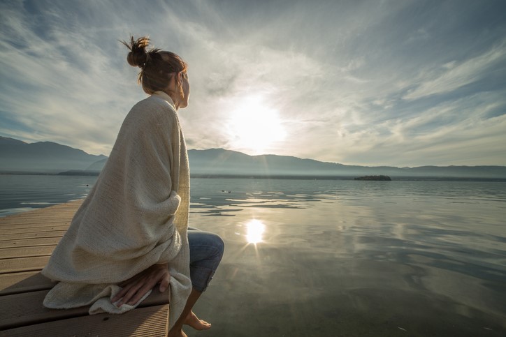 A woman wrapped in a blanket sits on a pier overlooking a calm lake.