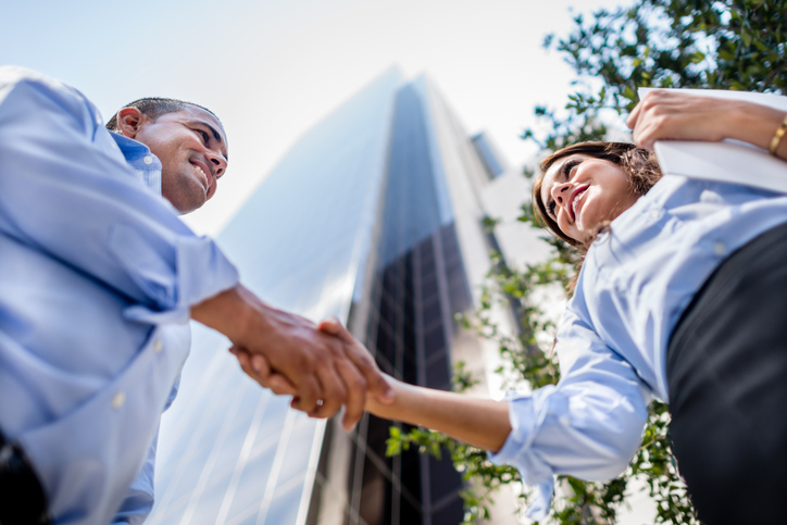 Business professionals shaking hands in front of a building. The camera is beneath the handshake, pointing up.