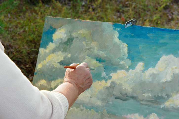 A senior artist works outside, painting a blue sky and clouds. 