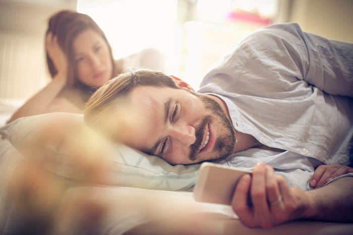 Man looks at phone in bed while his partner watches from a distance.