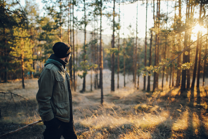 A man dressed for cold weather watches the sun set through the trees.