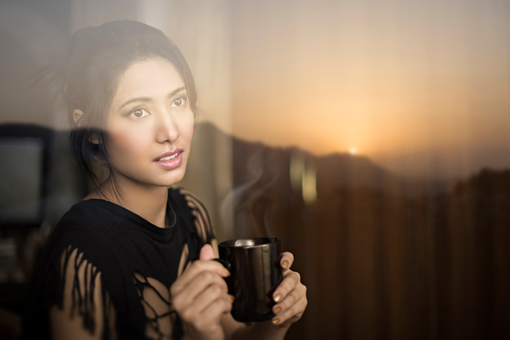A woman in a dark room watches the sunrise out her window.