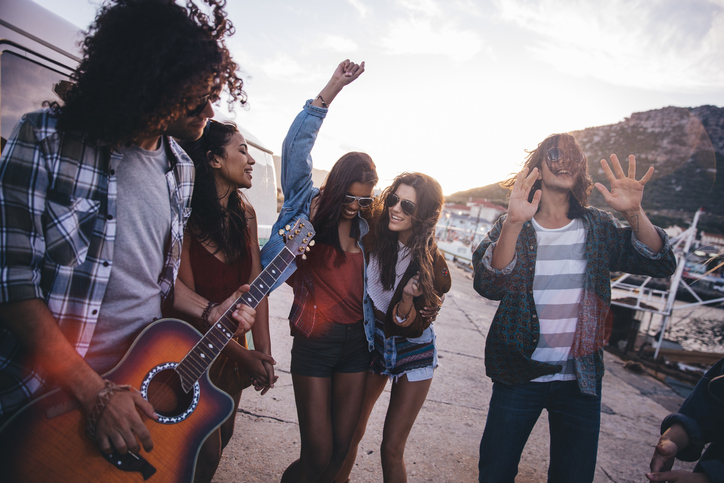 A group of friends walks around a harbor. The person in front has a guitar.