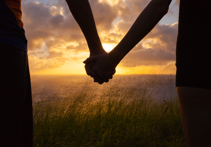 Close-up of two people clasping hands before a sunset.