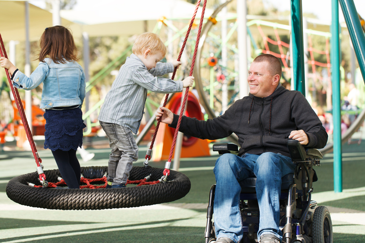 A father in a wheelchair helps his young kids use a playground course.
