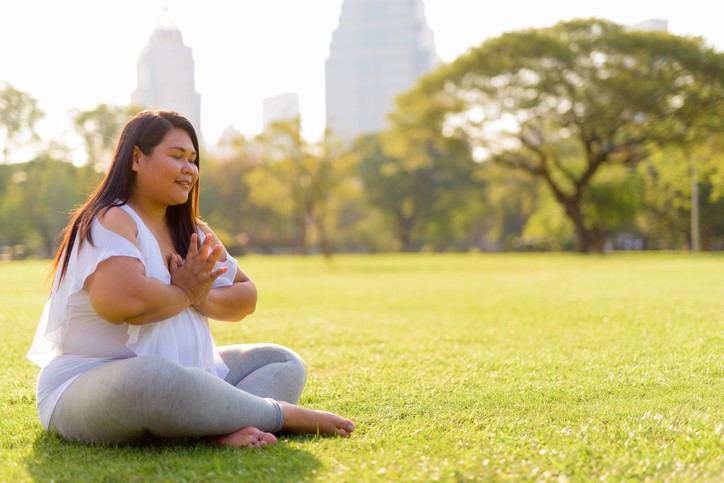 A young woman meditates on an open, sunny lawn. There are silhouettes of buildings on the horizon.