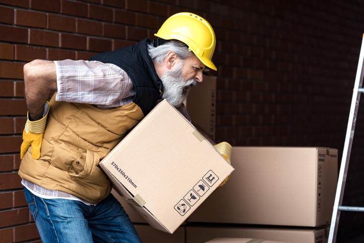 Factory employee in a hard hat pauses his work to clutch his lower back. 