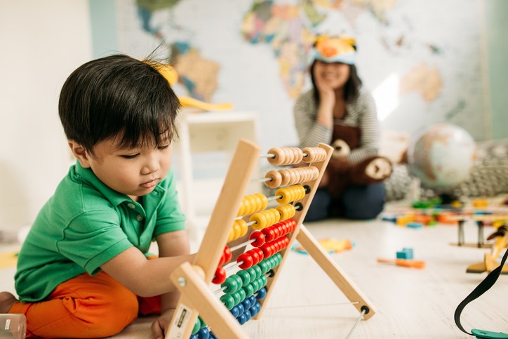 A toddler plays with an abacus while his mother watches from across the room.