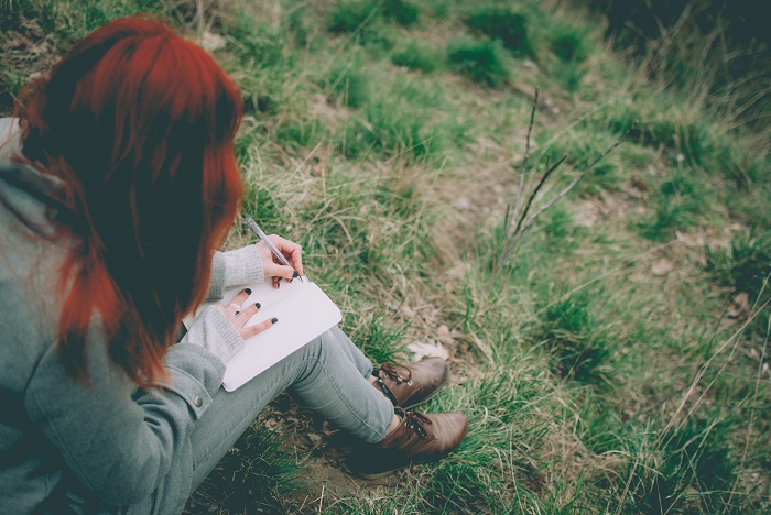 Girl with red hair sitting on some grass, writing in a journal