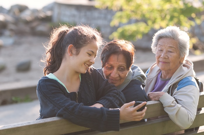 One girl sitting on a bench with two senior women, showing them her phone.