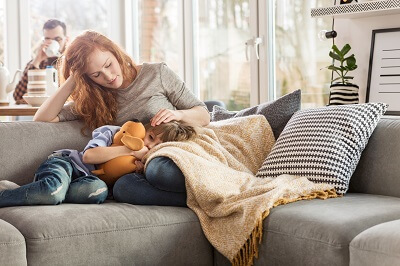 Mother sits with unhappy son on the couch while distant father drinks coffee in the background