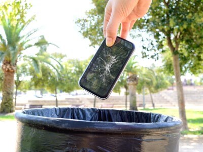 A hand is about to throw a cracked smartphone in a public trash can.