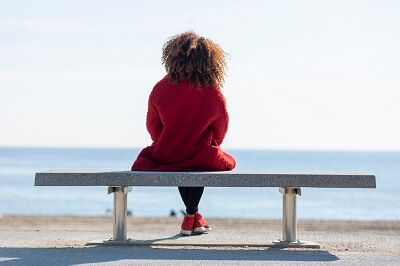 Woman in red jacket sitting on bench near ocean, alone