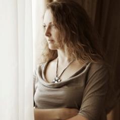 A woman stands hugging herself, looking out a window.