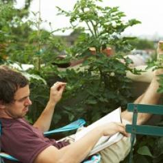 A man, his feet kicked up on a bacolny, reads a book near plants.