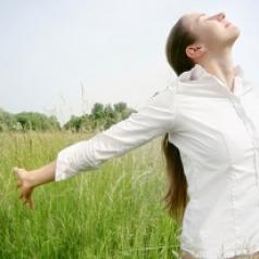 A woman stands in a grassy field and streatches her arms out while taking a deep breath.