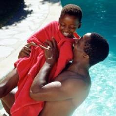 African-American father and son next to a pool in the sunshine. The father is lifting his son, who is wrapped in a red towel, up into the air in celebration. Both are smiling happily.