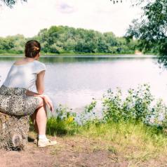 Person in skirt with hair pulled back sits on tree stump at lakeside looking over water
