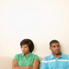 Couple sitting in front of white wall, arms crossed, looking away from each other as if frustrated
