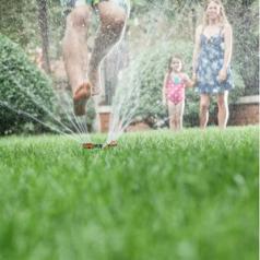 A father jumps over a sprinkler as his wife and daughter watch.