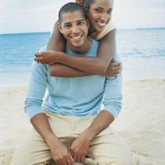 Young black couple on sandy beach. Both are smiling and kneeling on the sand. She has her arms wrapped around his shoulders from behind him.