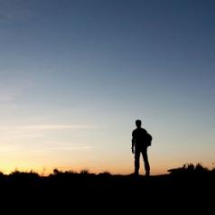 A silhouette of a man looks out at the sunrise on the summit of a hill. 