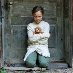 Child crouching in door of abandoned house with teddy bear