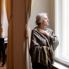 Senior adult wearing elegant brocade wrap with white hair in a clip stands at window looking outside