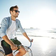 adult with facial hair, short hair, rides bike along shore smiling