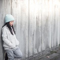 Preteen with long hair under light blue hat stands leaning back against fence, looking thoughtful
